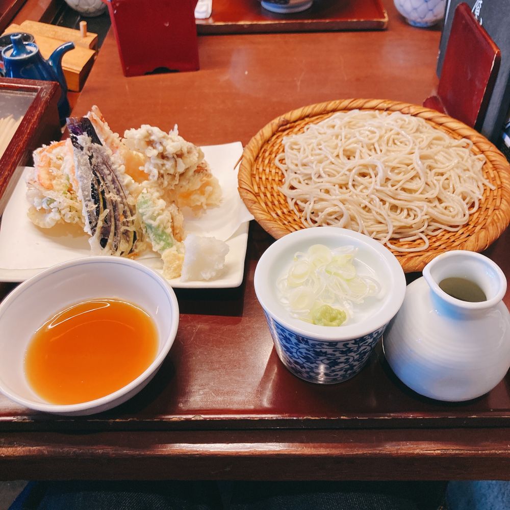 赤茶色のお盆。手前側にてんつゆ、蓋の部分にネギとわさびが乗ったそばつゆ入れ、そばつゆの入ったとっくり型の器がある。その奥に数種類の天ぷらが盛られた角皿、ざるそばがある。