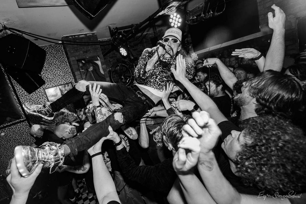 A dynamic black-and-white photo captures the raw energy of Norwegian punk band The Good The Bad The Zugly performing live at Enga, a tightly packed indoor club. Center stage—though physically above it—is vocalist Ivar, crowd surfing atop a sea of raised hands. He’s fully dressed, wearing a patterned jacket, sunglasses, and a hat, gripping a microphone as he leans forward mid-performance. The crowd beneath him is dense and animated, arms outstretched to support his weight, creating a powerful sense of unity and chaos. Stage lights beam through the smoky air, casting dramatic highlights and shadows across the scene. The background reveals typical club gear—speakers, lighting rigs, and cables—framing the moment in gritty realism. The photo freezes a peak of punk adrenaline, with Ivar suspended above the crowd like a rebellious icon, immersed in the noise, sweat, and connection of live music.