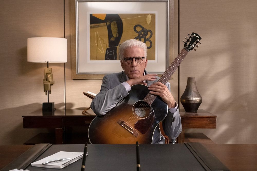 Michael from The Good Place, sitting at his desk and leaning on his guitar (which he is bad at playing)