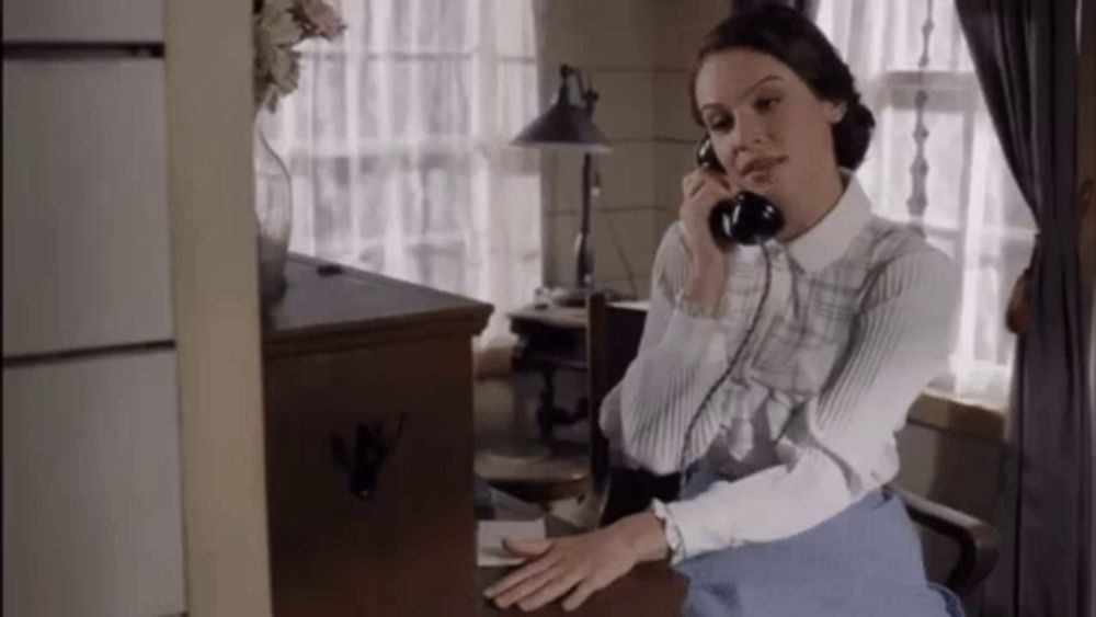 a woman is talking on a telephone while sitting at a desk in a room .