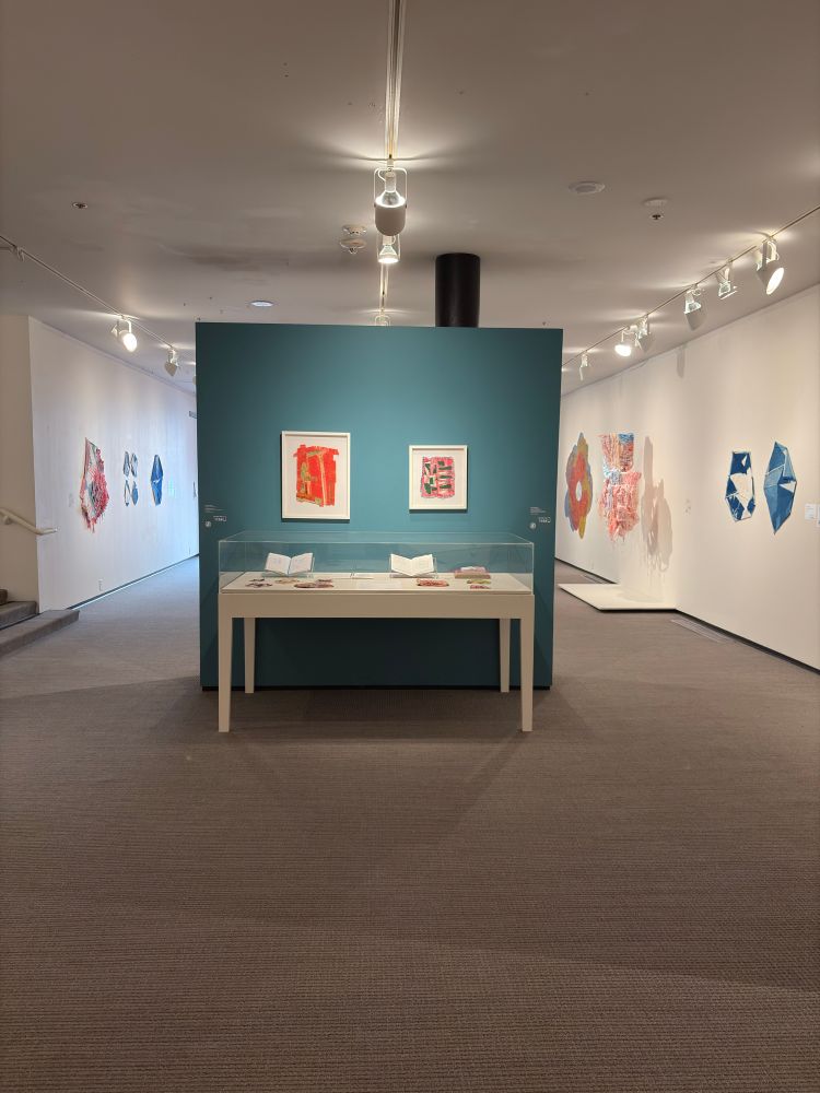 Framed drawings hang on a divider wall in a white cube gallery. The wall is painted a teal blue. In front of the wall is a vitrine with sketchbooks and other small works.
