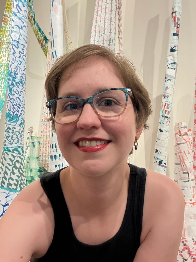A white woman with short blonde hair, glasses, and red lipstick smiles in front of painted paper scroll towers