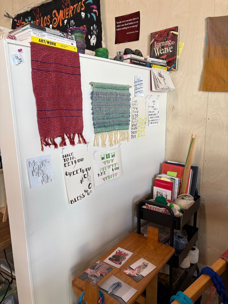 A corner of a studio with weavings and text-based drawings (concrete poetry?) on the walls. Books are stacked on top of the short wall and on a cart. On a wooden loom bench sit four cards of my artworks.