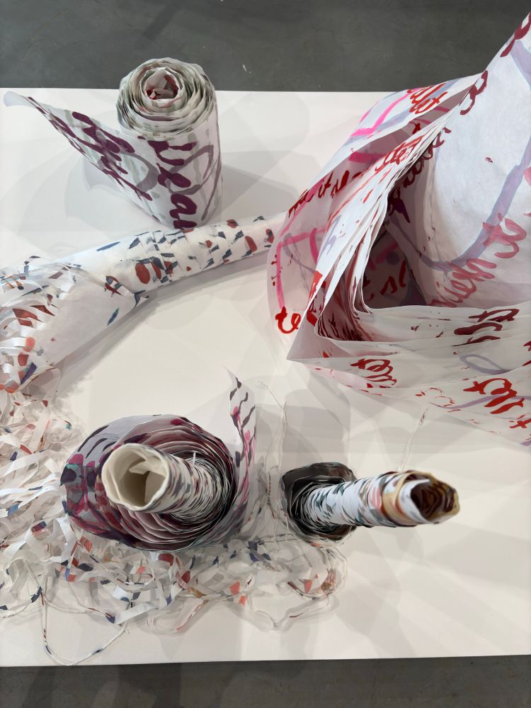 Painted scrolls of paper shot from above on a wide white pedestal. One is laid on its side and is split into fringe, with the ends spun into yarn.
