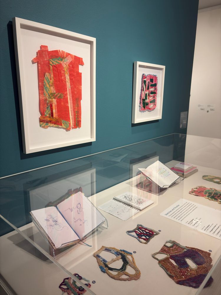 Two framed works on paper hang on a teal blue wall above a vitrine. The vitrine contains open sketchbooks and organic shaped crochet “drawings” (psychedelic doilies, if you will)