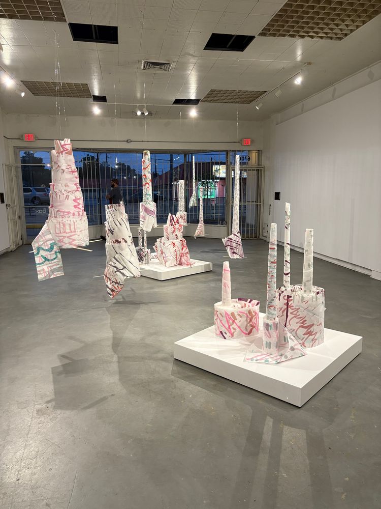 A white box gallery shot from the back of the room. “Spindles” of paper scrolls are suspended from the ceiling are spread throughout the space. Repeated forms of paper rolls spiral upwards and unspool a bit on two white pedestals low to the ground.