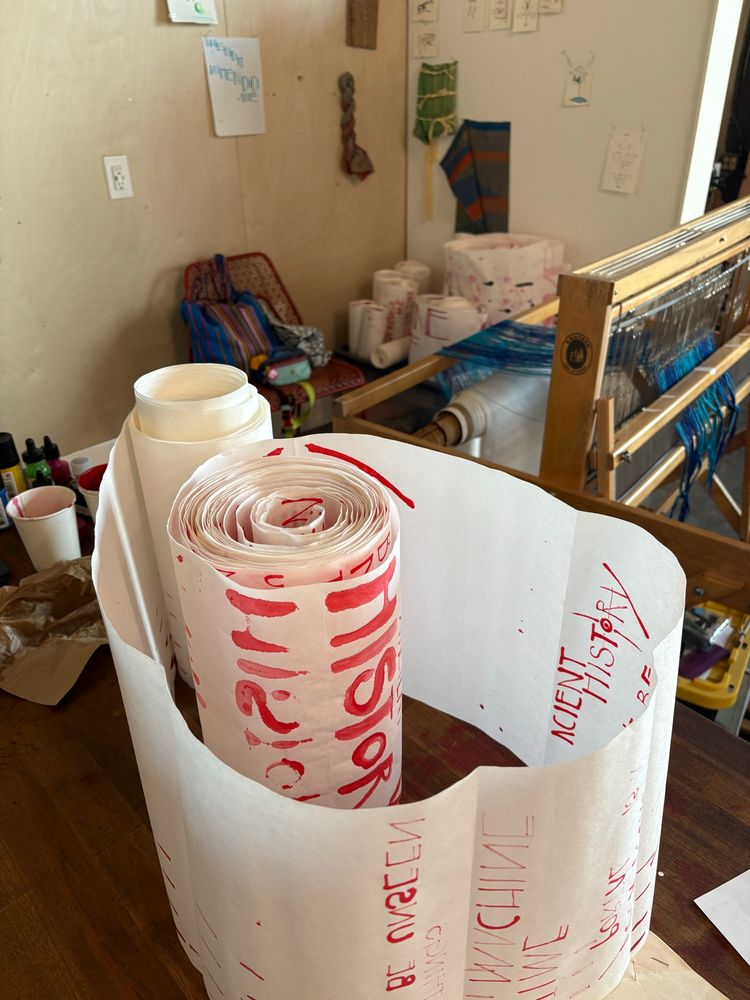 A scroll of white paper with red writing stands on end on a desk with both sides curled and a big swoop unfurled between them. Behind is a loom and more scrolls.