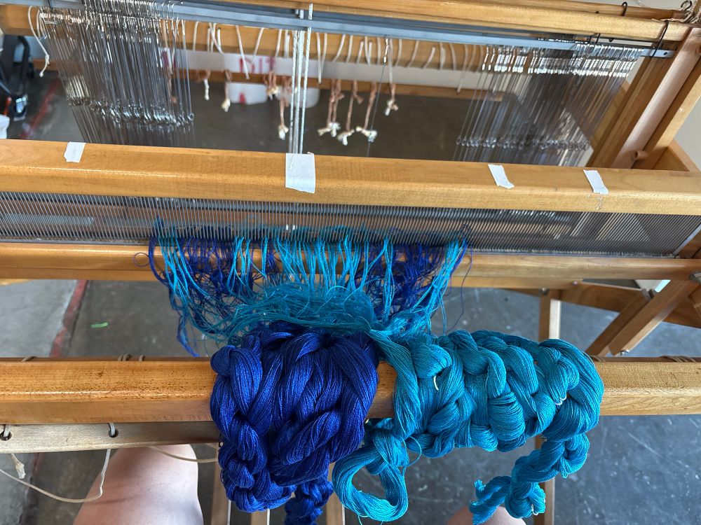 Dark and light blue threads are chained and wrapped around the front beam of a floor loom. The ends have been threaded through the comb-like part of the loom, called the reed.