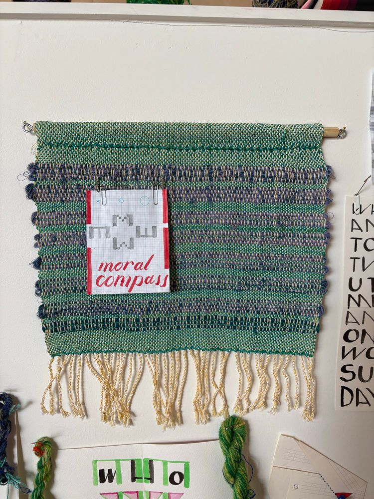 My studio wall detail shot: a weaving in mostly greens with yellow-white fringe hangs on a dowel. Pinned to the side like a brooch on its chest is a drawing on a grid. The drawing in red ink white negative space and graphite is both graphic and textual: MEWE reads the compass, offset and weighed down (smooshed). A calligraphic script reads “moral compass”. Around the weaving hand other text-drawings concrete poems and hanks of green handspun wool yarn.