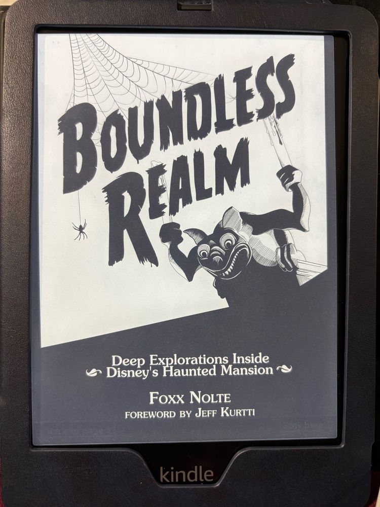 A Kindle Paperwhjte in a navy blue case showing the cover of Boundless Realm: Deep Explorations inside Disney’s Haunted Mansion by Foxx Nolte