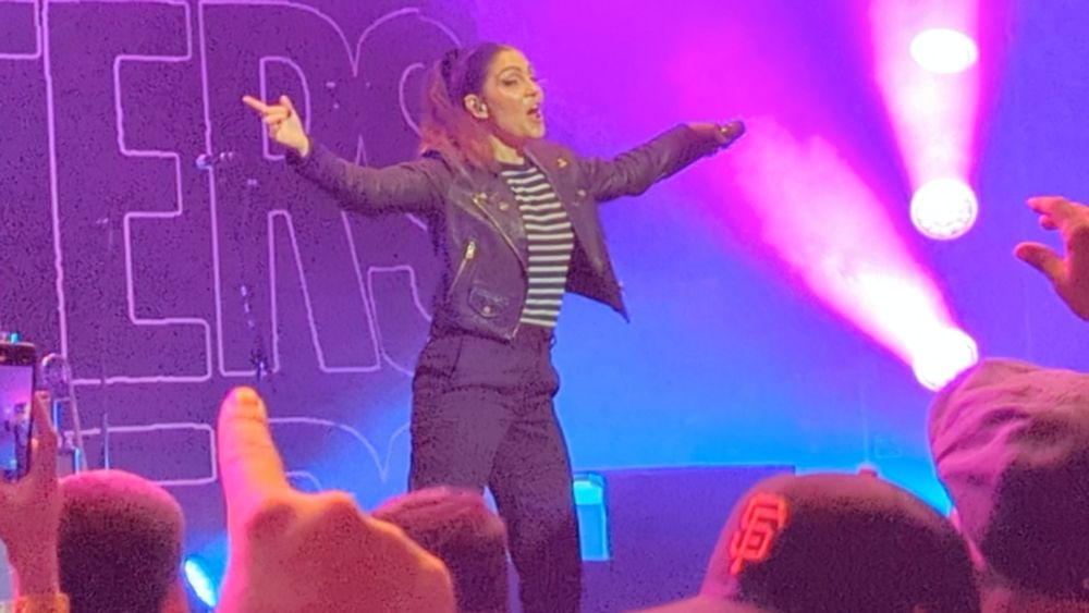 Aimee from the interrupters during their show at The Masonic in San Francisco. She has a ponytail, black pants, and a black coat over a black and white horizontal striped shirt. Behind her is a backdrop with the bands name shown multiple times and multiple purple stage lights.