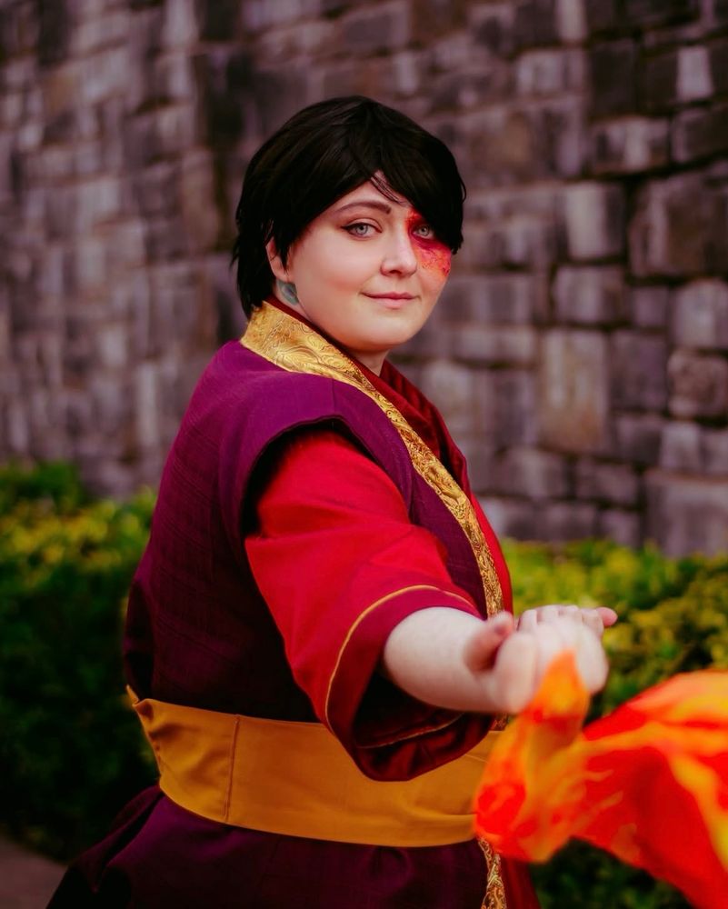 Misha is cosplay as Zuko from Avatar the Last Airbender standing in a pose with one fist out and red fabric makes it look like she’s firebending 