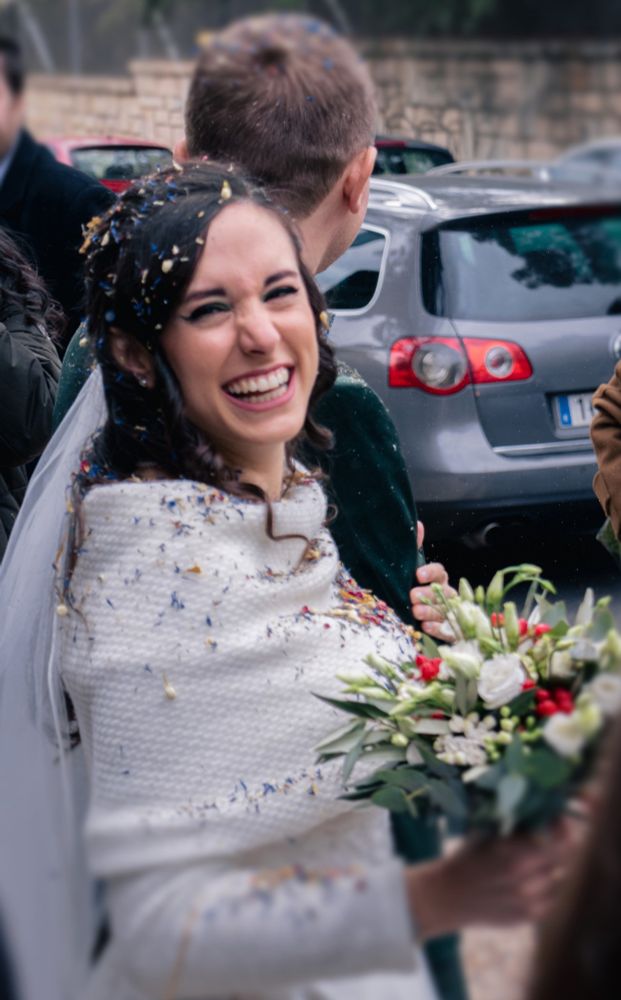 A newly wed couple holding arms. Groom looking away, and bride in a long-sleeved white dress smiling to the camera holding a bouquet of winter flowers.