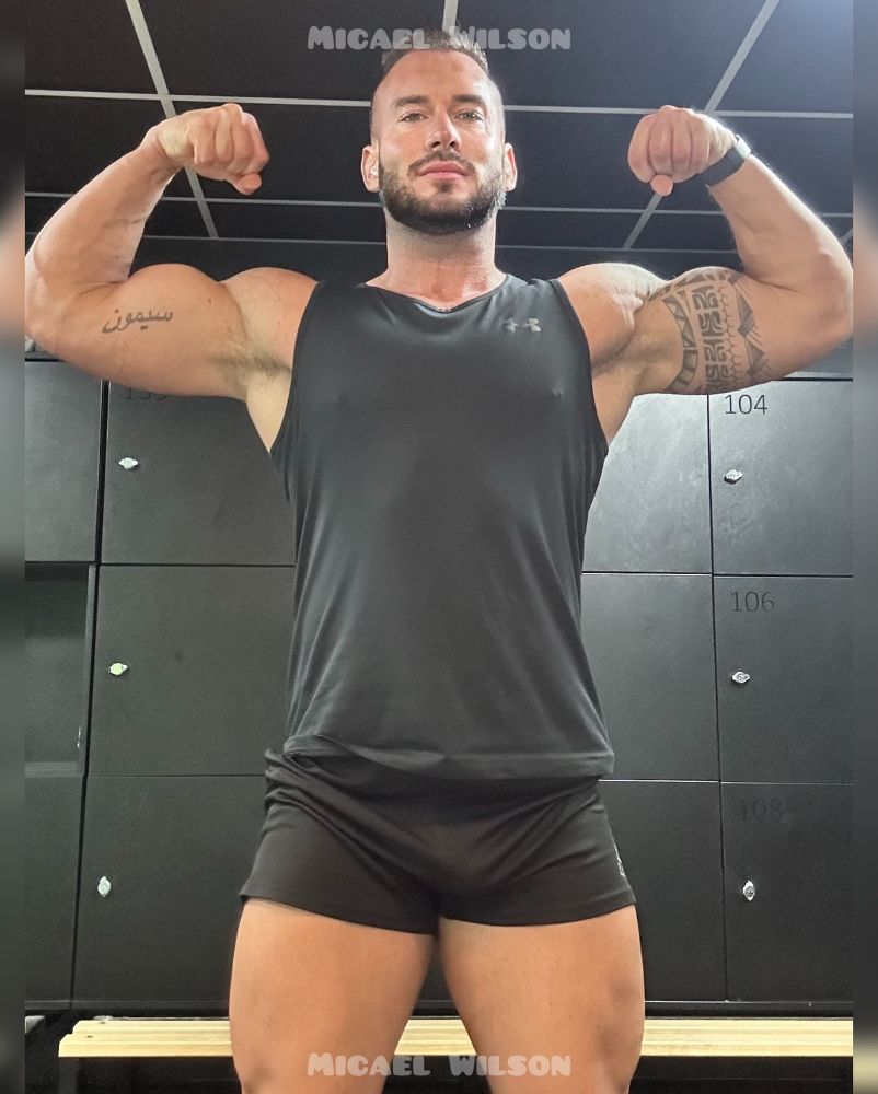 Flexing in the mirror with both biceps up and flexed wearing a black under armour fest and black shorts as a bench and black lockers in the background