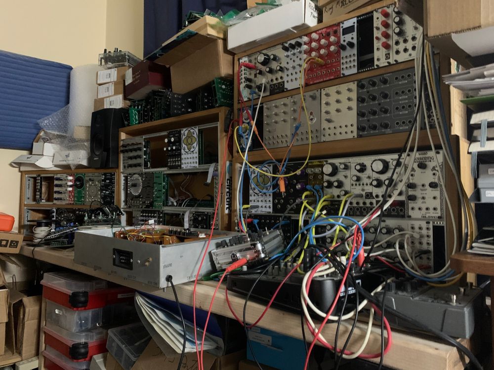 several racks of eurorack synthesizer modules, audio electronics, kit boxes, parts storage boxes and cables running every which way