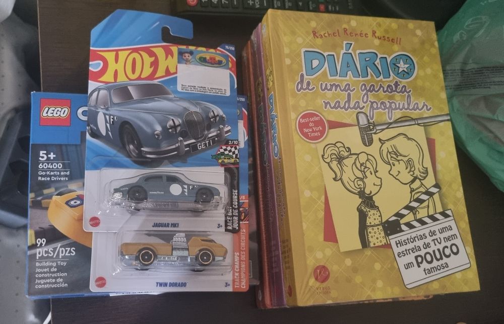 Um caixa pequena de lego que está embaixo de dois carrinhos da hotweels (um azul e um dourado) na caixa. 
Livro amarelo do diário de uma garota nada popular em cima de outros dois livros. Na capa do livro tem o título o desenho de uma menina de cabelo preso em um rabo de cavalo alto de perfil olhando para um menino de cabelo médio também de perfil, ambos sorriem sem mostrar os dentes.
