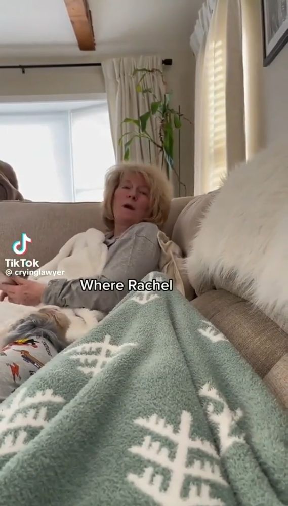 Mulher branca e loira sentada em um sofá debaixo de cobertas. A imagem é vista do ângulo lateral. Na imagem tem o texto "where rachel" (onde rachel)