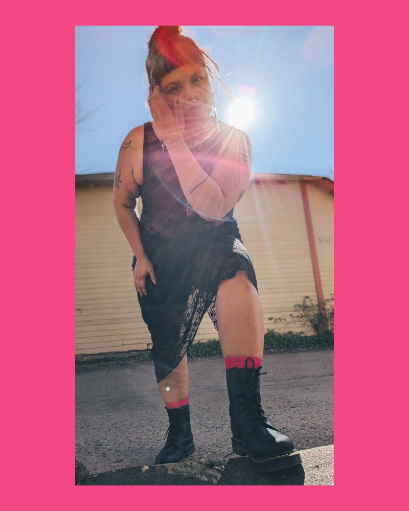 Bruja leans toward the camera the sun creating a halo over their shoulder. They are in a long lace dress, black leather boots and pink boot socks. They are gently pushing hair away from their face and gazing intensely at the camera.