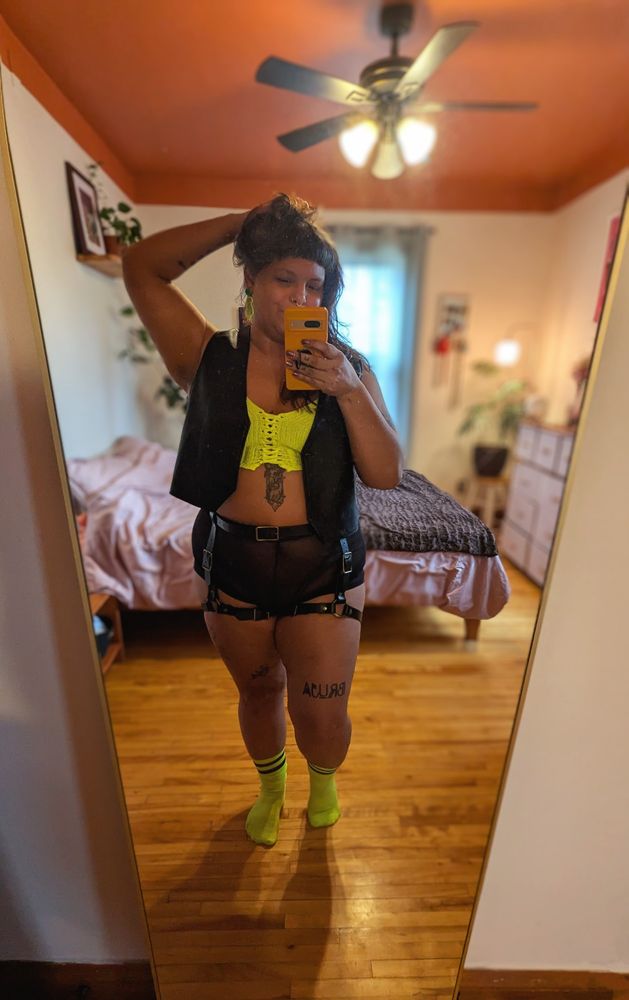 Bruja poses for a mirror selfie in a leather vest and harness over mesh boxers and high visibility neon crocheted top & tube socks.