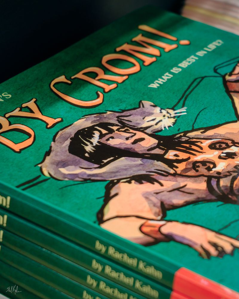 A stack of books titled By Crom! with Conan the Barbarian on the cover chilling with a cat on a couch.