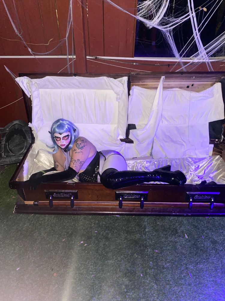 Draggedy Anne with giant alien eyes sitting in a coffin dressed in blue hair, elf ears, a purple romper, and black gloves and boots. 