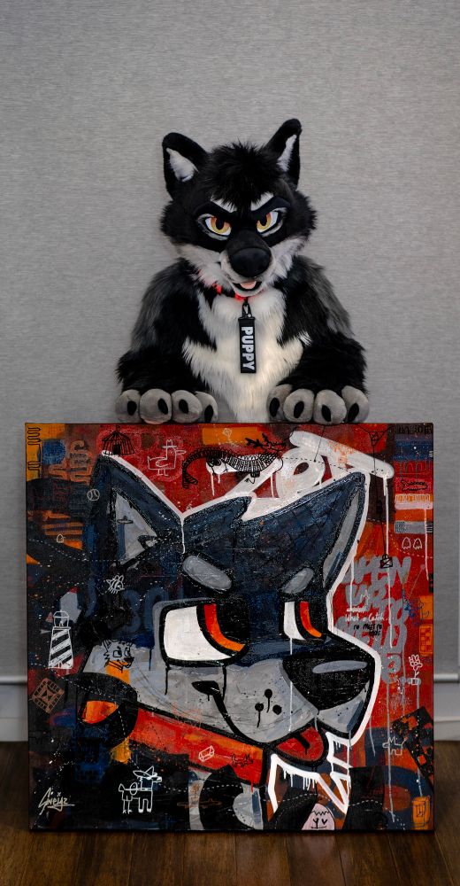 A black, white, and grey wolf fursuit poses behind a colorful painting. The fursuit has warm orange eyes, a black muzzle, and a tag on the collar that reads 'PUPPY'. The painting in front of them is a bold, street art-style portrait of the same character, featuring exaggerated eyes, sharp teeth, and a red, black, and orange background filled with graffiti-like symbols and text."
