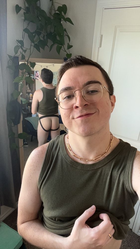 Selfie of myself wearing a green tank top, rose gold necklace, and eye glasses with my butt appearing in the mirror reflection behind 