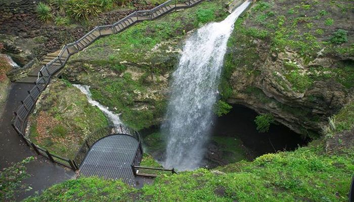 A waterfall flowing into a huge sinkhole, covered with vegetation. A walkway has been built around the sinkhole, leading to a viewing platform