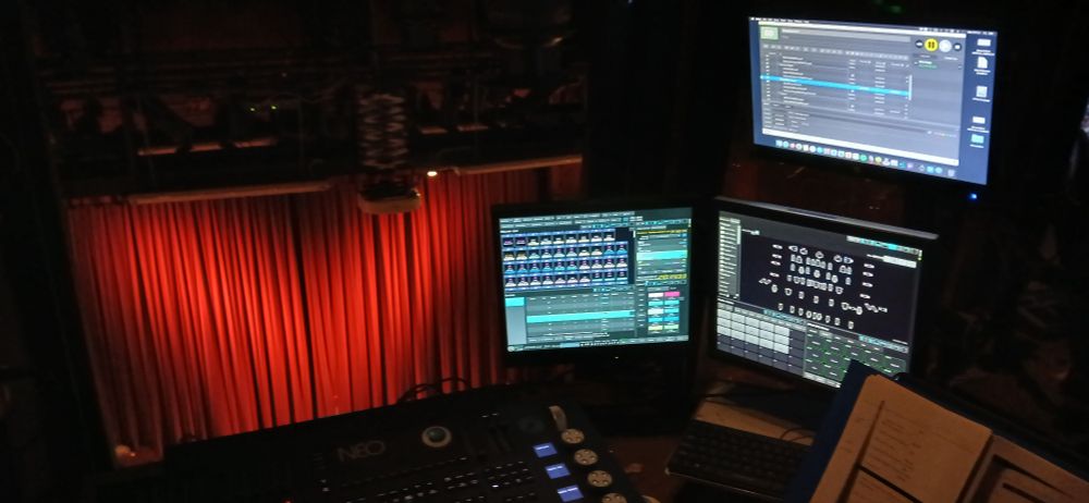 View from the control room of a theatre. In the foreground are a lighting console, a script, and monitors showing lighting and AV software. In the background, red curtains are closed over the stage