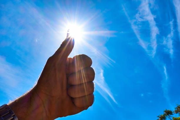 Photo of a man's hand in a thumbs up, in front of a blue sky with the sun partially covered by the thumb