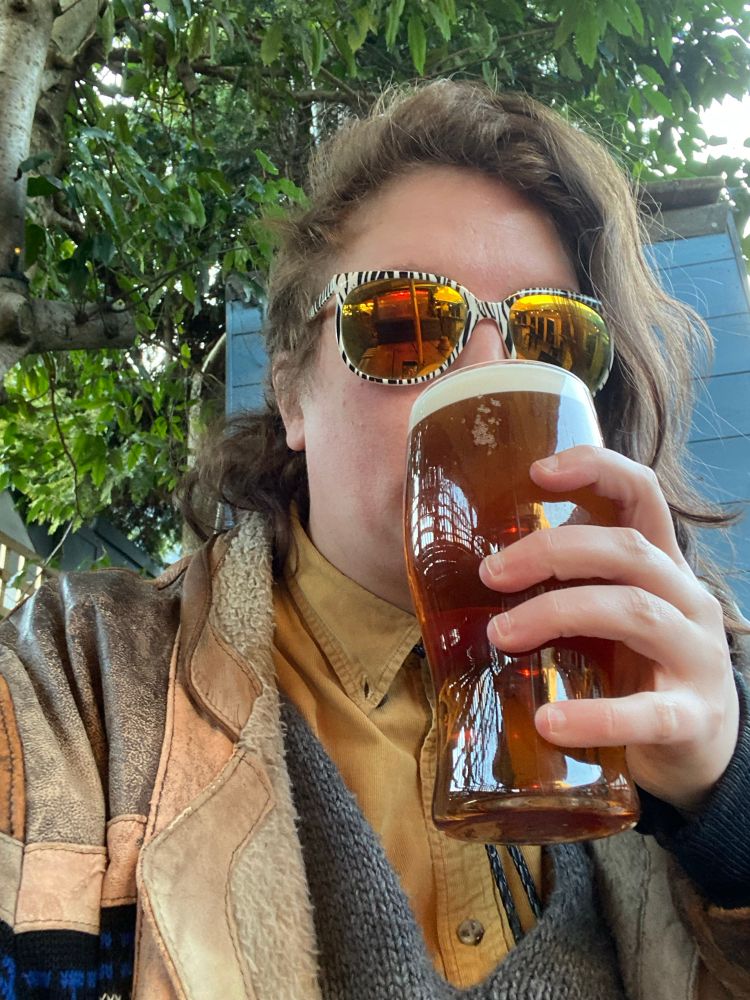 a person (the owner of the account, thorn) with an asymmetrical haircut, wearing a mustard yellow button up shirt, brown leather jacket, a knitted grey poncho and some reflective sunglasses. they are sat in a beer garden, with a decent amount of foliage nearby. they have a full pint glass raised to their mouth and whimsy in their heart.