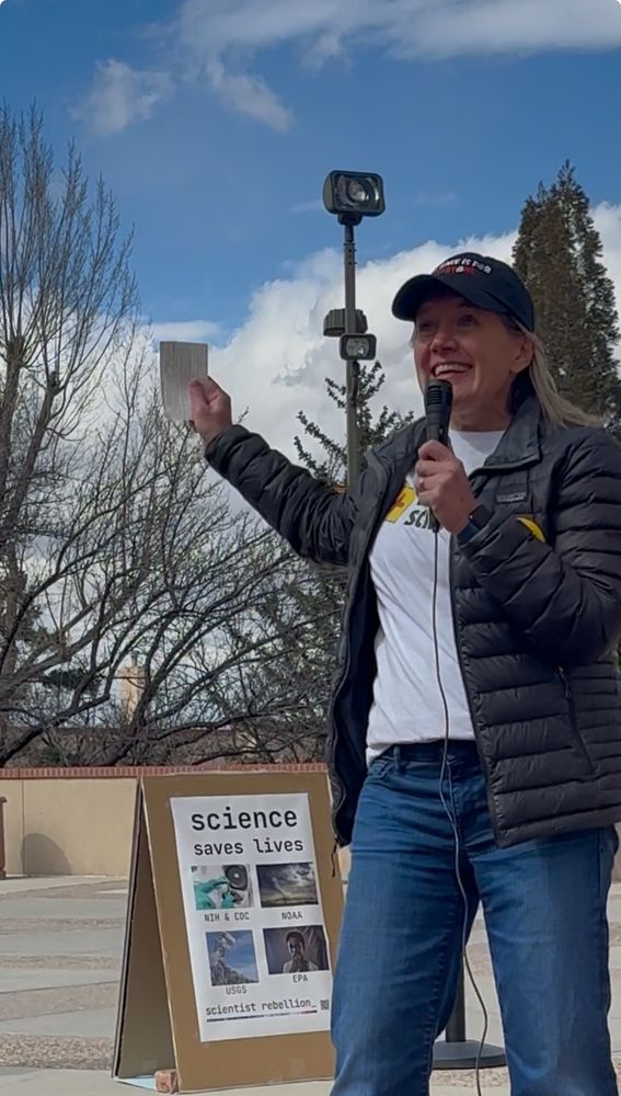 Person speaking with microphone at rally. Background sign says “science saves lives”