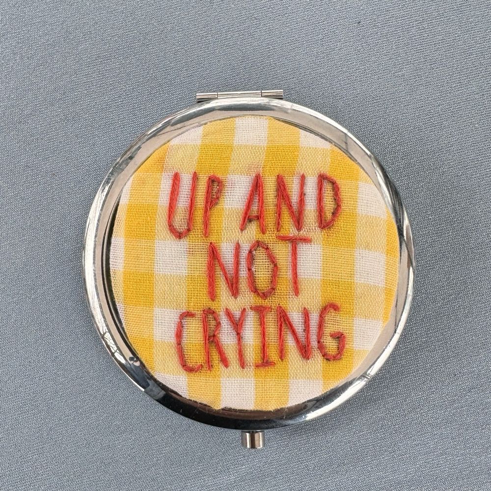 A closeup of a silver pill box laying flat. The top says “up and not crying” in orange capital letters on yellow and white plaid fabric