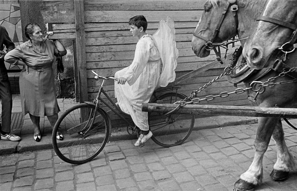 Un enfant enfant déguisé en angelot, lors d'une parade, descend une pente à vélo dans un village, suivi par un attelage de chevaux.