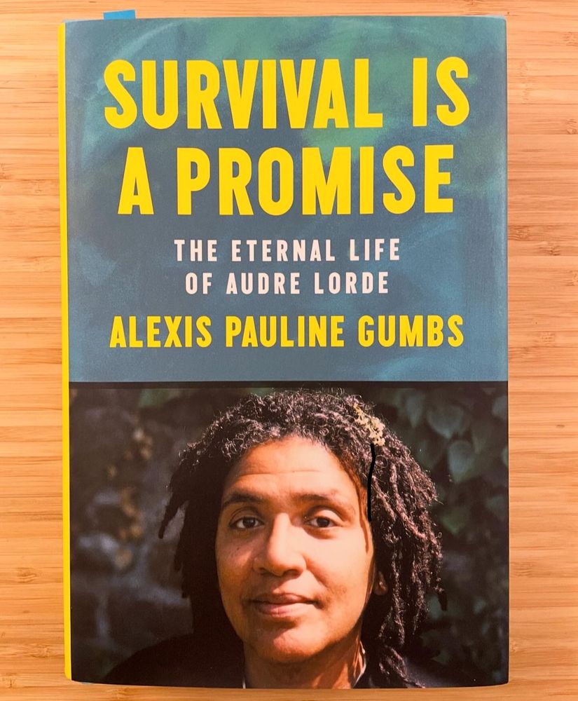 A hard cover copy of Alexis Pauline Gumbs’ biography of Audre Lord on a desk—“Survival is a Promise: The Eternal Life of Audre Lorde.” Its cover features Gumbs’ favorite picture of Lorde, one of her looking directly into the camera, centered, with a slight, knowing smile/resolve on her face.