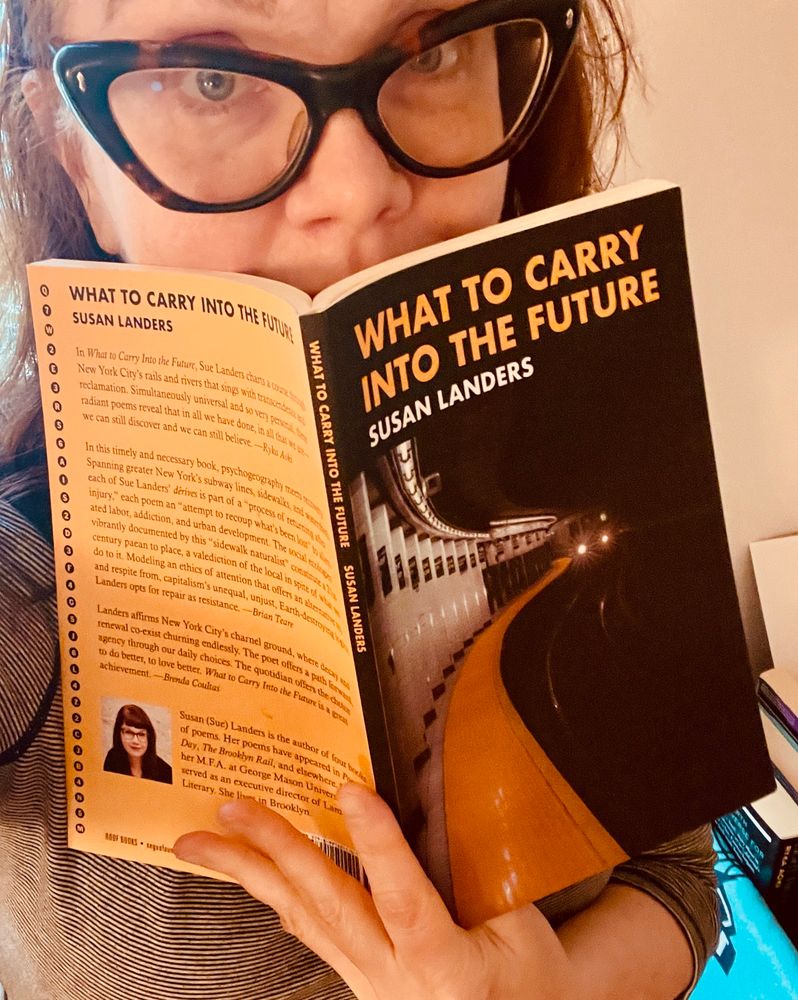 Person with cat eye glasses holds up the book What to Carry into the Future—poems that emerged from riding every subway line end to end, and featuring Astor Place on its cover and an author photo on the back.