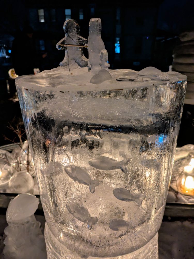 A luminary made of ice depicting two figures ice fishing with ice fish swimming inside the luminary