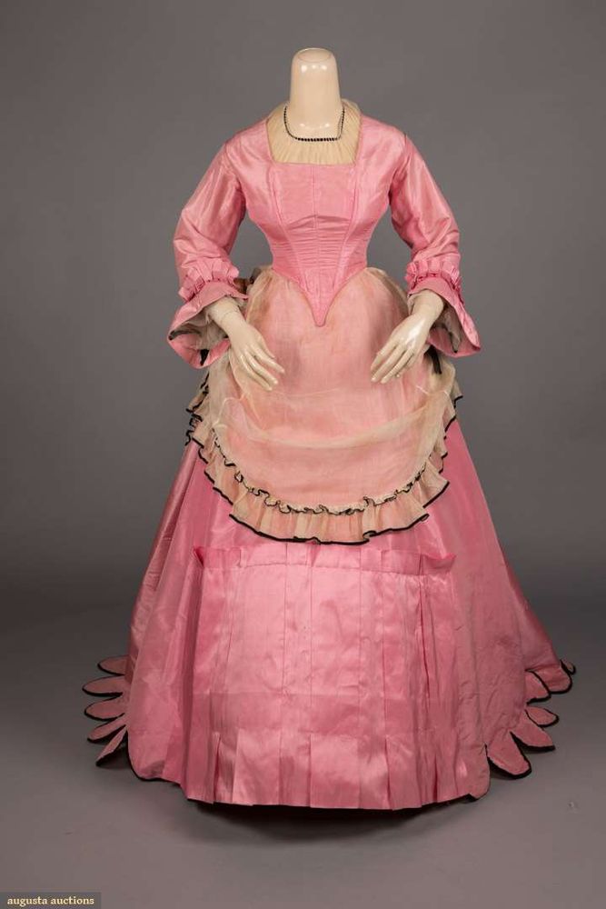 Description for all views, which is 4 different angles. A bubblegum pink silk taffeta dress on a mannequin, long sleeves, square neckline, pleated trim on sleeve hems. Bodice ends in point in front and has laced closure in back. The skirt front seems to be pleated. The back half of skirt has scalloped edge at hem. 


Sheer cream (I suspect it is patina and was white) over skirt forming in an apron-like manner in front, and also long in back, but with lifted sides with black velvet ribbons at sides. This piece is trimmed with black velvet ribbon.
