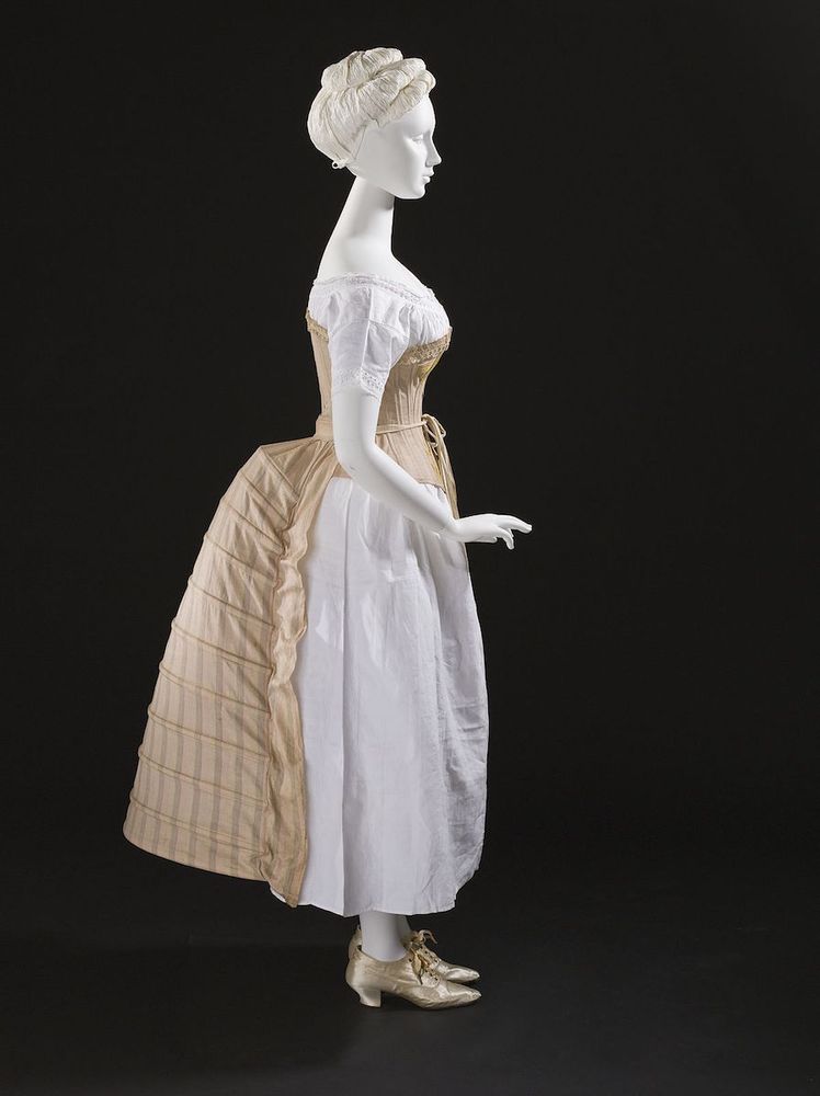 Mannequin wearing a beige colored corset, a white long chemise and beige striped bustle. 