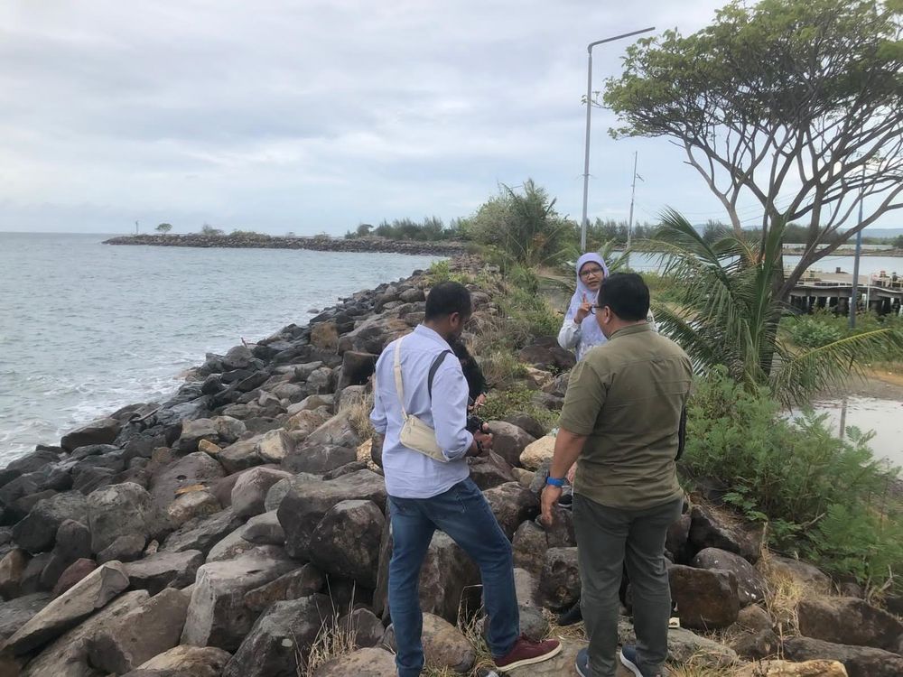 Four people stand on a seawall in Indonesia. It is a low rubble wall, with the sea on one side and a port on the other.
