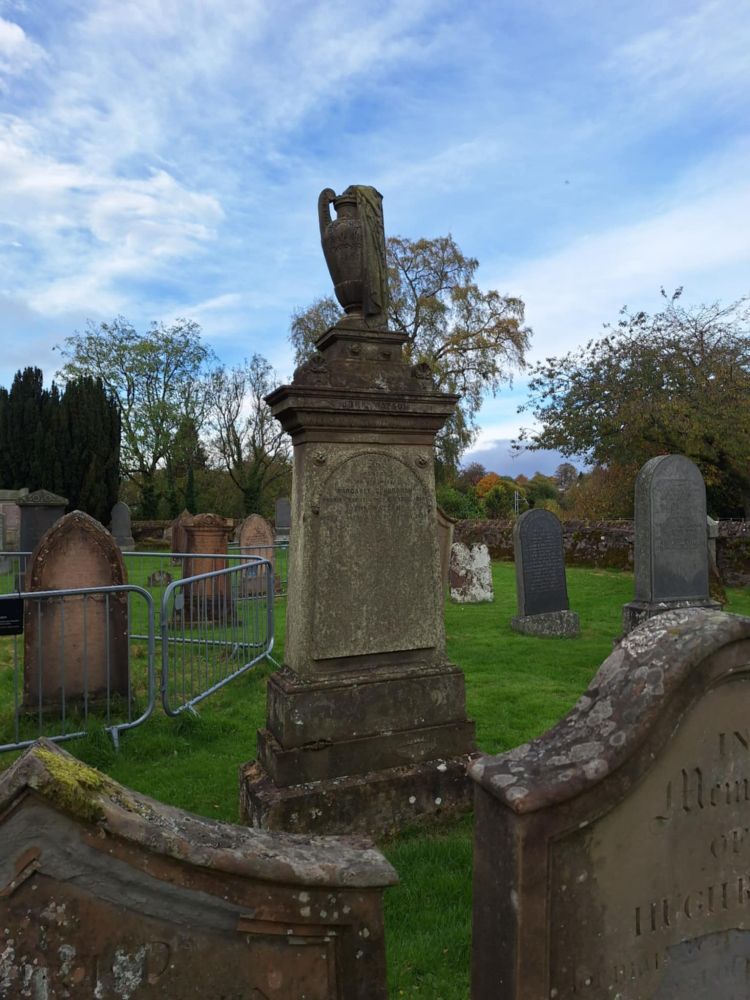 Large sandstone headstone with draped urn on top, surrounded by other headstones and fencing. Trees in background and some blue sky at last!