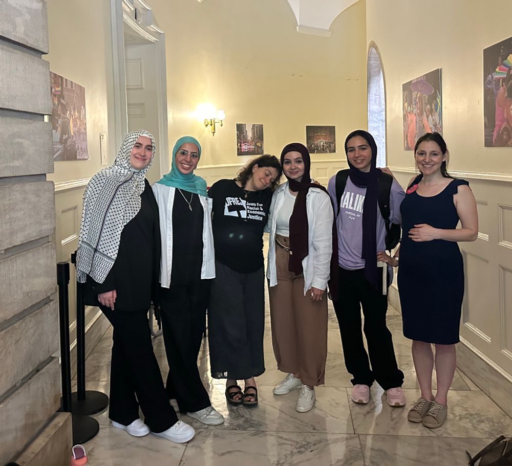 Photo of four Muslim women and two Jewish women smiling, standing together in a marble hallway. 