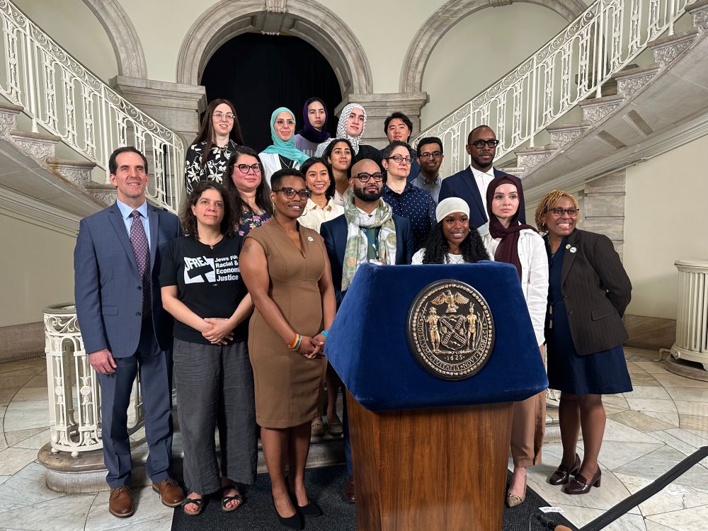 Photo of a diverse group of people standing behind a podium in City Hall. 