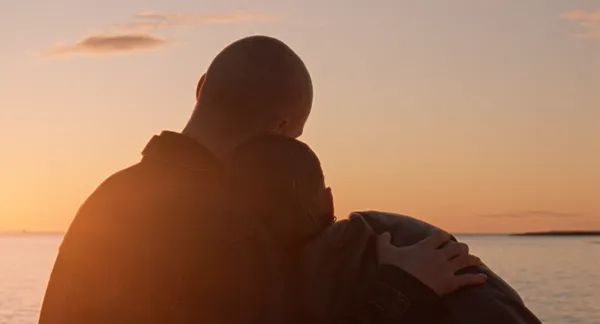 A still from the film When The Light Breaks. Two white people, one with a shaved head and the other with shoulder-length hair, sit on a clifftop, holding one another and watching a tangerine-coloured sunset.