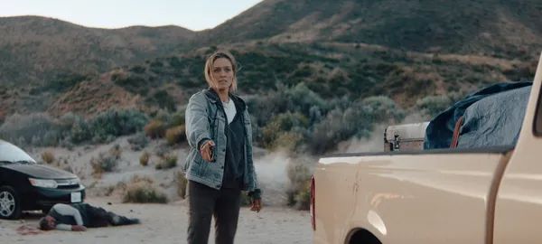 A still from the film Redux Redux. A white woman stands beside a truck in desert scrubland, aiming a gun. Behind her, another figure lies on the ground beside a car, and there is blood in the dirt.