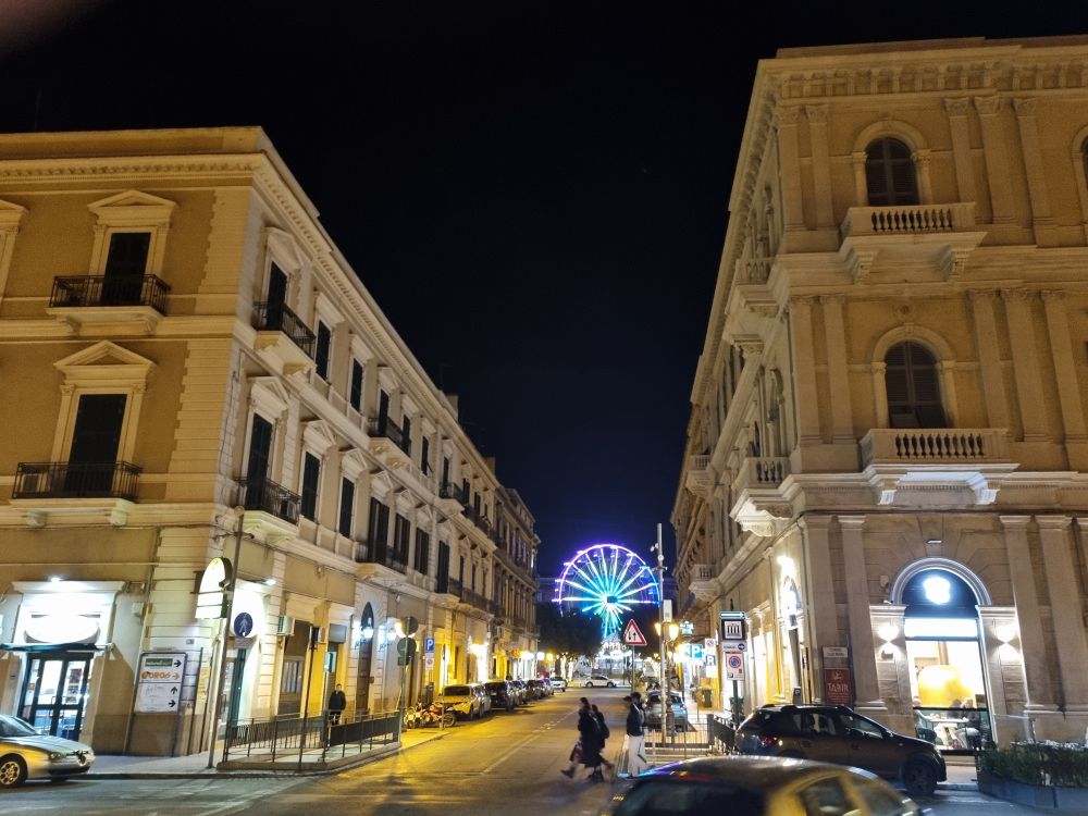 eine Strasse ist von zwei dreigeschossigen sandfarbenen Häusern eingerahmt. Es ist Nacht, die Fensteröffnungen sind dunkel und am Ende der Strasse ist ein Riesenrad mit Lichtern zu sehen. Es leuchtet blau und türkis.