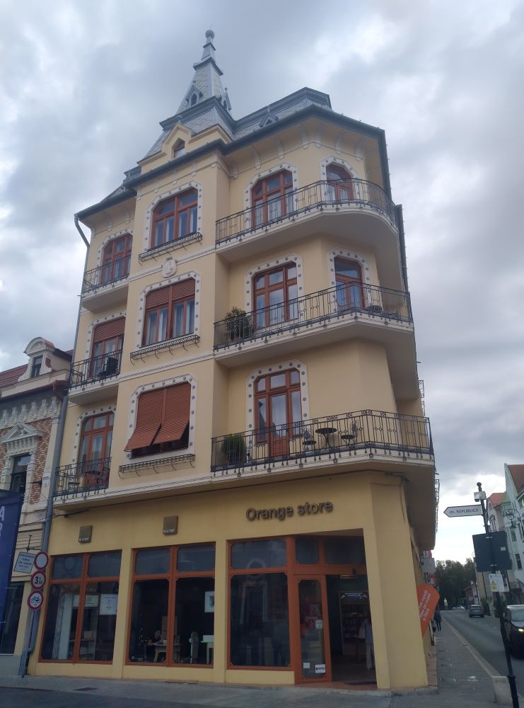 closeup view of the front side of a yellow thin mid rise building with 4 levels on the street corner in the center of a city, on the ground level there is a commercial space with the words Orange store written above the entrance, the three upper levels have balconies on bot sides of the front separated by a wall with a window between them, the roof is grey metal sheet with a tower like triangular shape structure rising up in the middle