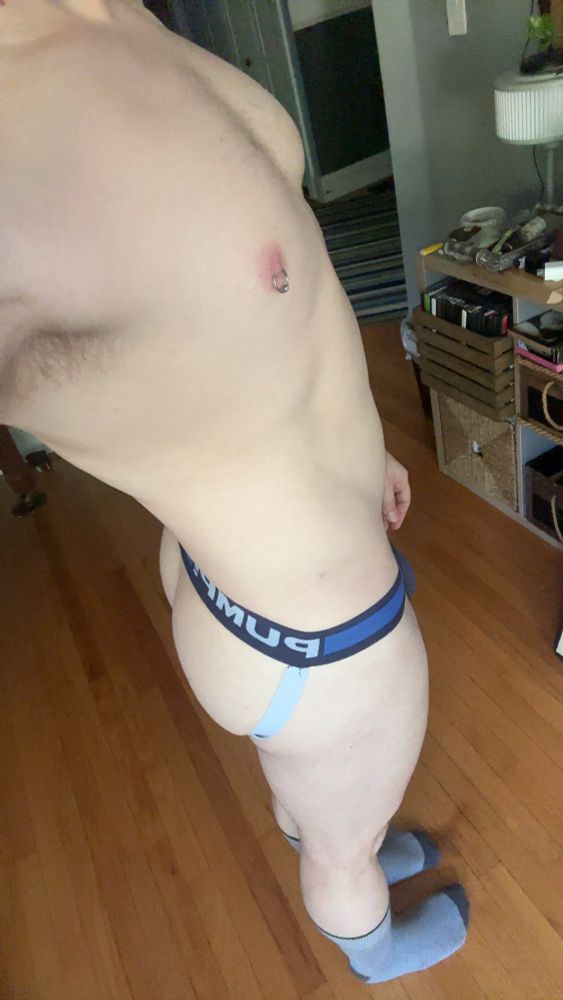 Selfie of a man with a nipple piercing from the neck down wearing a blue, black, and white jockstrap and gray socks 