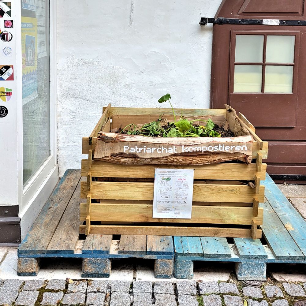 Foto einer Kompostkiste aus Holz mit dem Schriftzug "Patriachat kompostieren" und einer Anleitung, was kompostierbar ist und was nicht. In der Kiste befindet sich bereits Kompost und Erde, es wachsen ein paar kleine Zucchini-Pflanzen. 