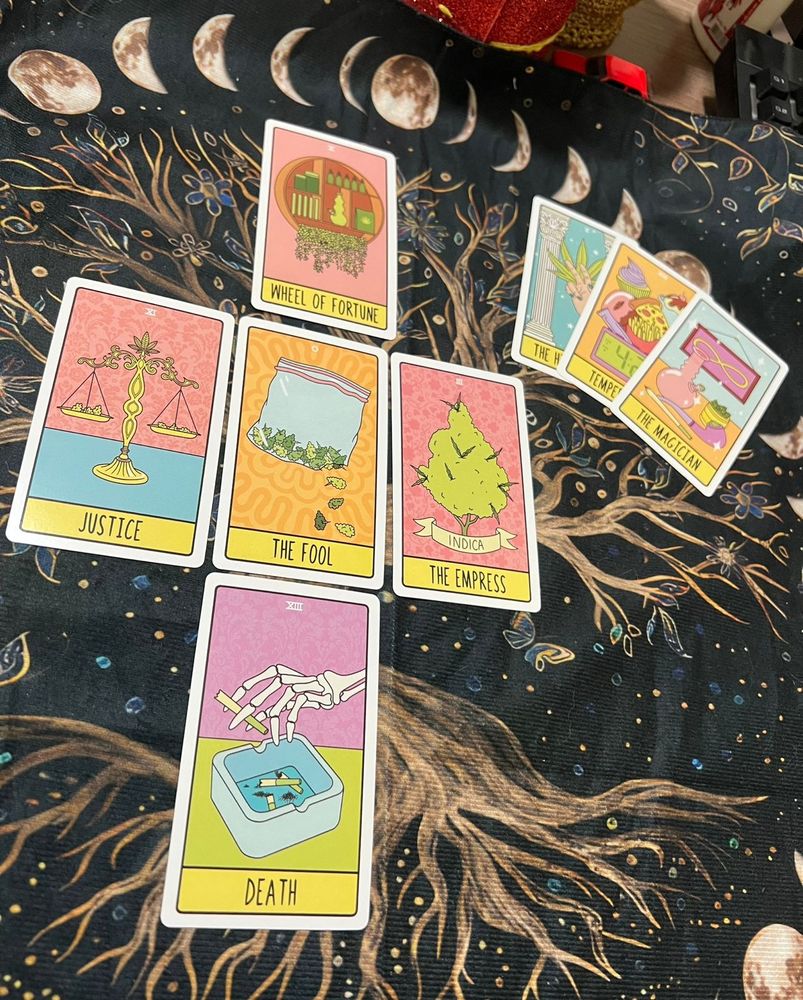 A imagem exibe um arranjo de cartas de tarô em jogo de peladán de um baralho temático, focado na cannabis, sobre um tecido escuro com elementos cósmicos e naturais.

casa 1 (Justiça) - XI: Apresenta uma balança dourada com suas bandejas cheias de flores de cannabis. O fundo da carta é rosa com um padrão sutil de damasco.
casa 2 (A Imperatriz) - III: A figura central é substituída por um grande bud de maconha, identificado com um banner escrito "INDICA". O fundo também é rosa.
casa 3 (Roda da Fortuna) - X: Ilustra um armário ou prateleira circular, sugerindo um espaço de cultivo ou armazenamento, com livros e plantas penduradas.
casa 4 (Morte) - XIII: Mostra um esqueleto segurando um beck quebrado, sobre uma bandeja cheia de pontas de becks apagados.
casa 5 (O Louco) - 0: Mostra um saco plástico ziplock cheio de flores de maconha, com algumas pétalas espalhadas. O fundo é em tons de laranja/amarelo.

trinca: Há fragmentos de O Hierofante, Temperança, que mostra ícones como cupcakes, pizza e o número 4:20, e O Mago.

O pano de fundo é predominantemente preto, o que faz as cartas coloridas se destacarem intensamente.
Na parte superior, são visíveis várias ilustrações das fases da lua (cheia, crescente), desenhadas em tons de marrom e bege.
A parte inferior e central é dominada pelo desenho estilizado de uma yggdrasil (árvore da vida) em tons de ocre, sugerindo conexão com a terra e os ciclos vitais.
Pequenos pontos de luz ou estrelas estão espalhados pelo fundo escuro, reforçando o ambiente místico.